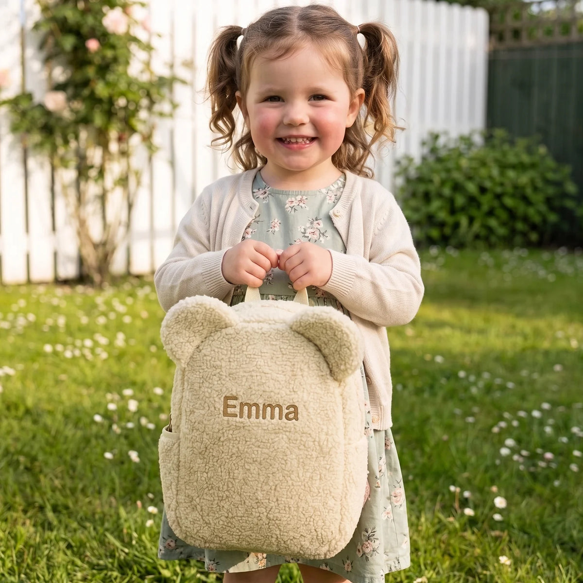 Petite fille tenant le sac à dos ourson beige avec le prénom « Emma » dans le jardin | market_france